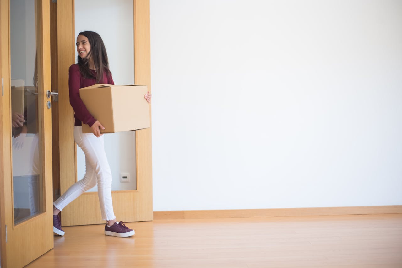 services-01 A woman enters a new apartment carrying a cardboard box, symbolizing moving in.