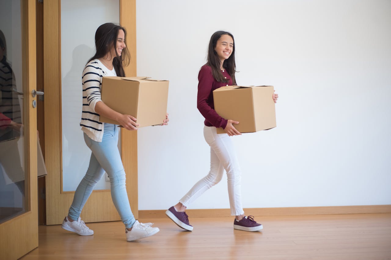 services-02 Two young women carrying cardboard boxes indoors, illustrating teamwork and moving in.
