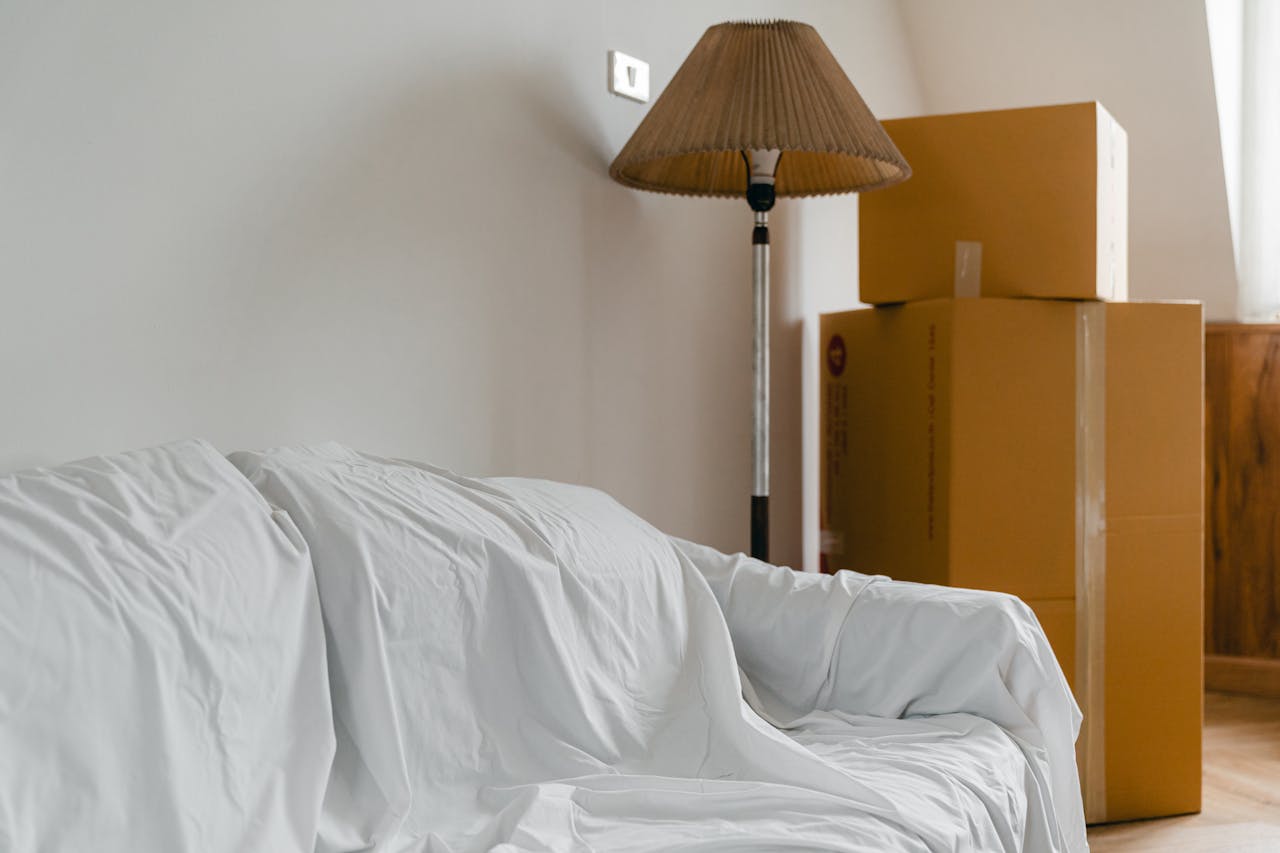 about-us Covered with sheet sofa and floor lamp and packed cardboard boxes in light spacious room of new contemporary house